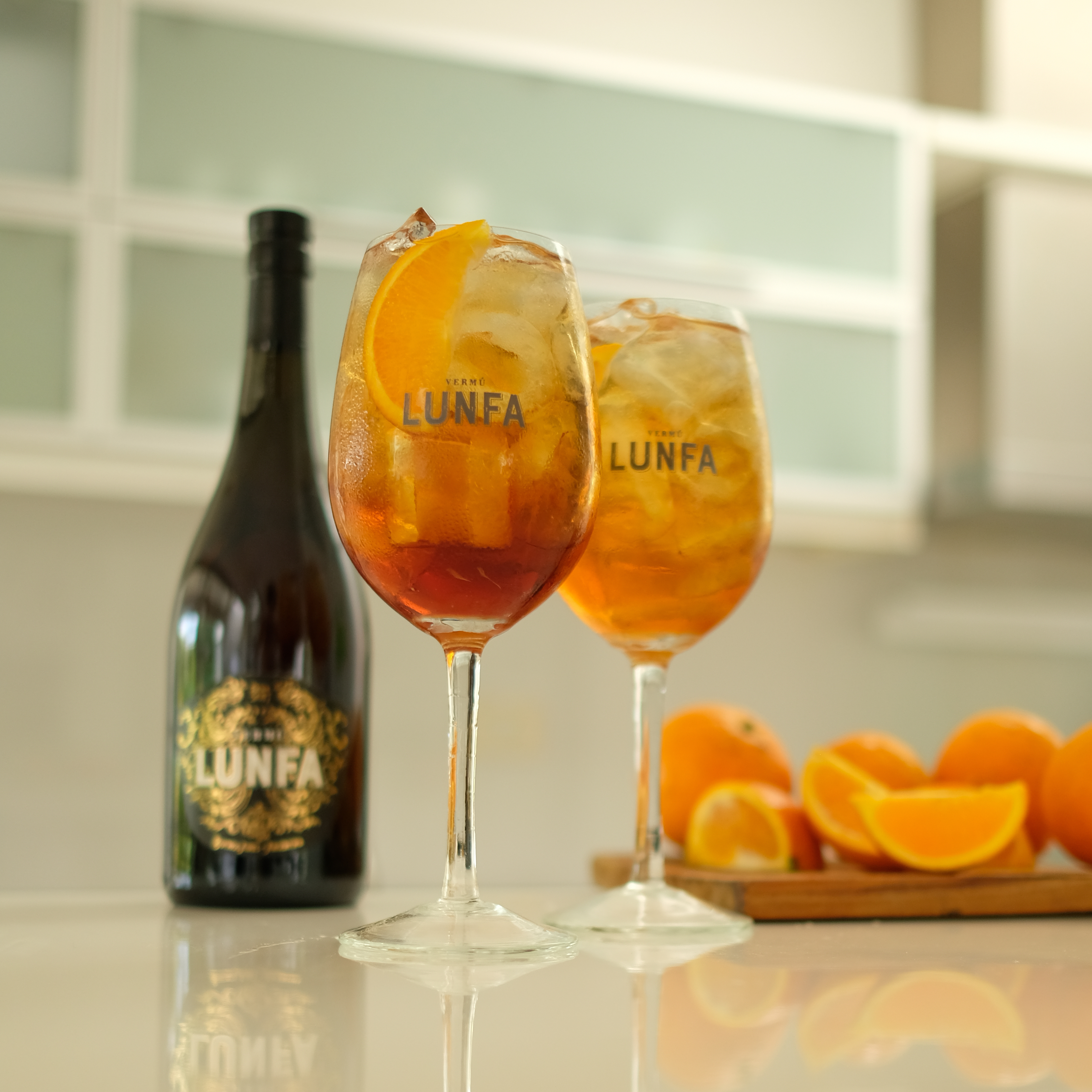 How to make Lunfa Spritz - two branded glasses filled with amber vermouth spritz, Vermú Lunfa bottle, and fresh oranges on white background